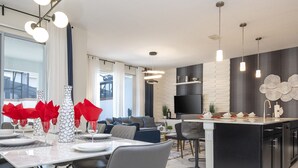 Een moderne keuken met een centraal eiland, een eetgedeelte met stoelen en een gedekte tafel, en een woonruimte met een bank en een televisie.