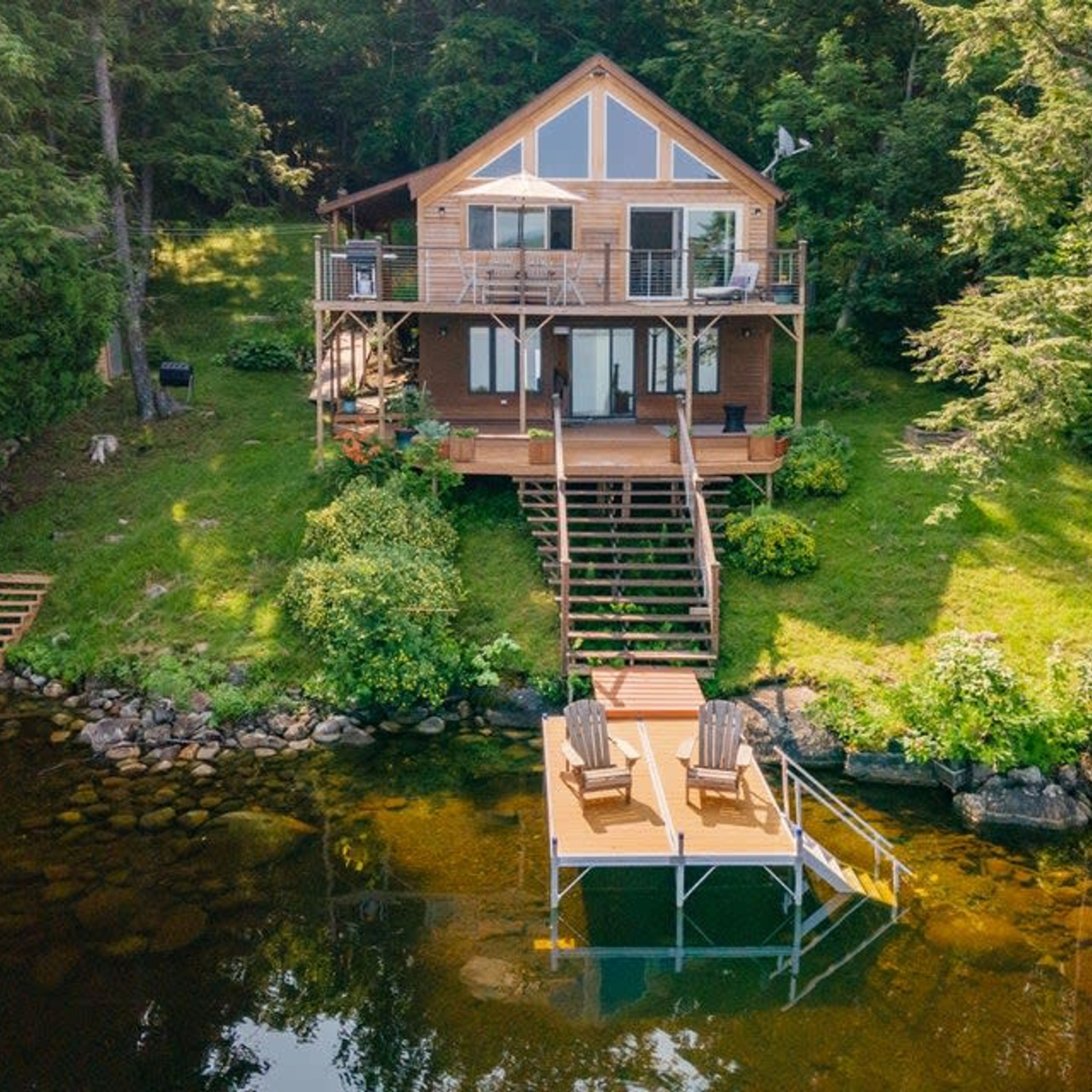 A two-story wooden cabin with a deck and a dock extending into a lake, surrounded by lush greenery.