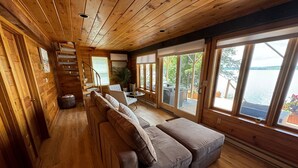 Ein Interieur einer Holzhütte mit einer Couch, einer Treppe und großen Fenstern mit Blick auf ein Gewässer.