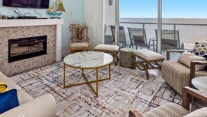 Una sala de estar moderna con chimenea, ventilador de techo, televisor grande y vista al océano a través de amplias ventanas.