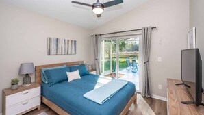 A bedroom with a bed, a nightstand, a lamp, a TV, and a view of a patio with a table and chairs. - Swiss 49 - Lake Denise (Clermont)
