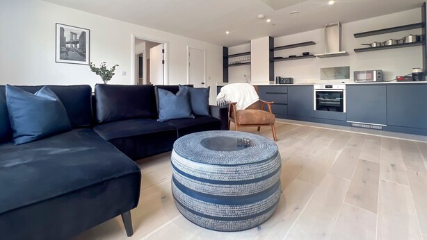 A modern living room with a black sofa, a round ottoman, and a kitchen in the background. - Manchester Industrial by Casacity (Manchester)