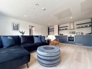 Un salon moderne avec un canapé noir, un pouf rond et une cuisine en arrière-plan.