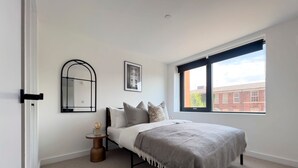 A bedroom with a bed, a round side table, a mirror, and a window with a view of buildings. - Manchester Industrial by Casacity (Manchester)