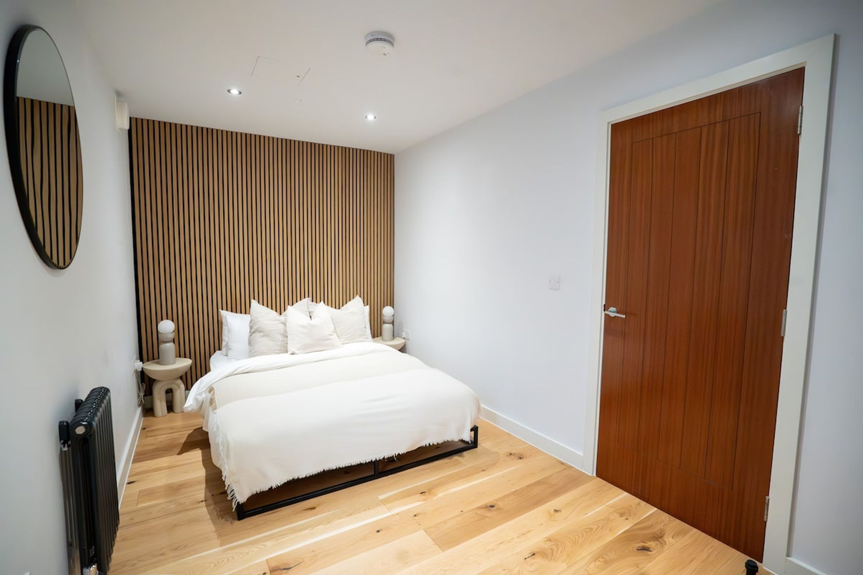 Een moderne slaapkamer met een houten vloer, een bed met wit beddengoed, een houten hoofdeinde, een ronde spiegel, een nachtkastje en een houten deur.