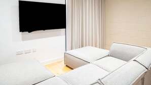Un salon moderne avec un canapé d’angle, un téléviseur à écran plat fixé au mur et une fenêtre avec des rideaux.