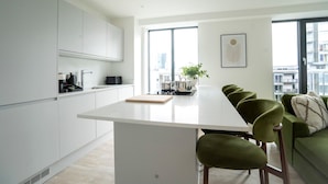 A modern kitchen with white cabinets, a central island, and a dining area with green upholstered chairs. - Organika Suite by Casacity (Salford)