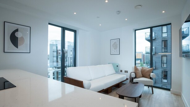 A modern living room with a white sofa, a coffee table, and a dining table. There are large windows offering a view of the city. - Alabaster Suite by Casacity (Salford)