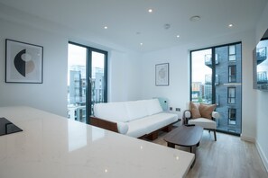 Una sala de estar moderna con un sofá blanco, una mesa de centro y una mesa de comedor. Amplias ventanas ofrecen vistas a la ciudad.