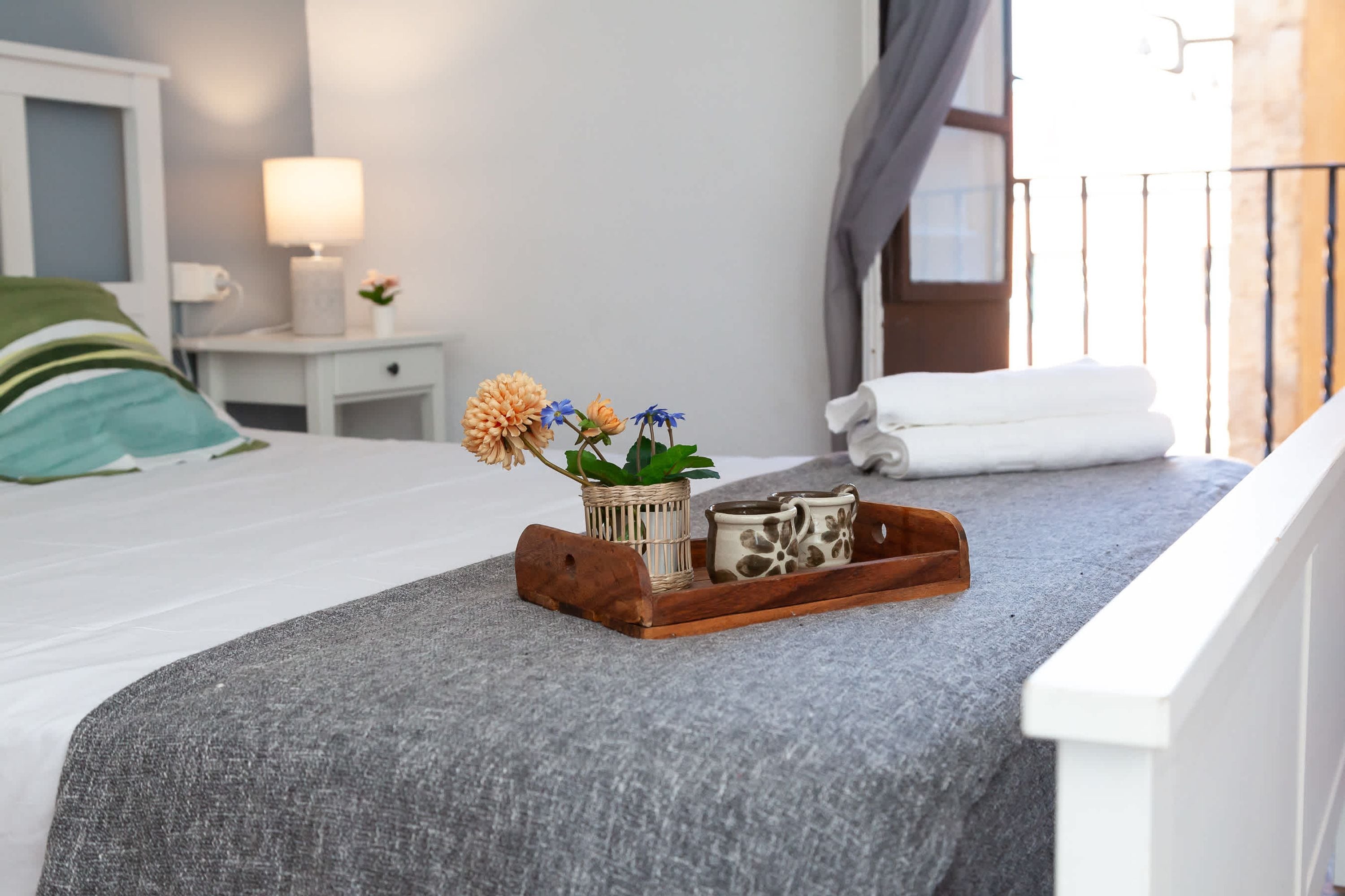 Una cama bien hecha con un edredón gris, una bandeja de madera con una planta y dos tazas, una mesita de noche con una lámpara, y una ventana con cortinas.