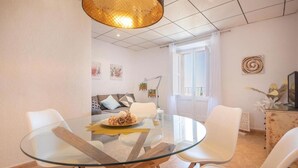 A modern living room with a glass dining table, white chairs, a sofa with cushions, a television, and a decorative wall piece. - Unik Vacation Rovellat (Tarragona)