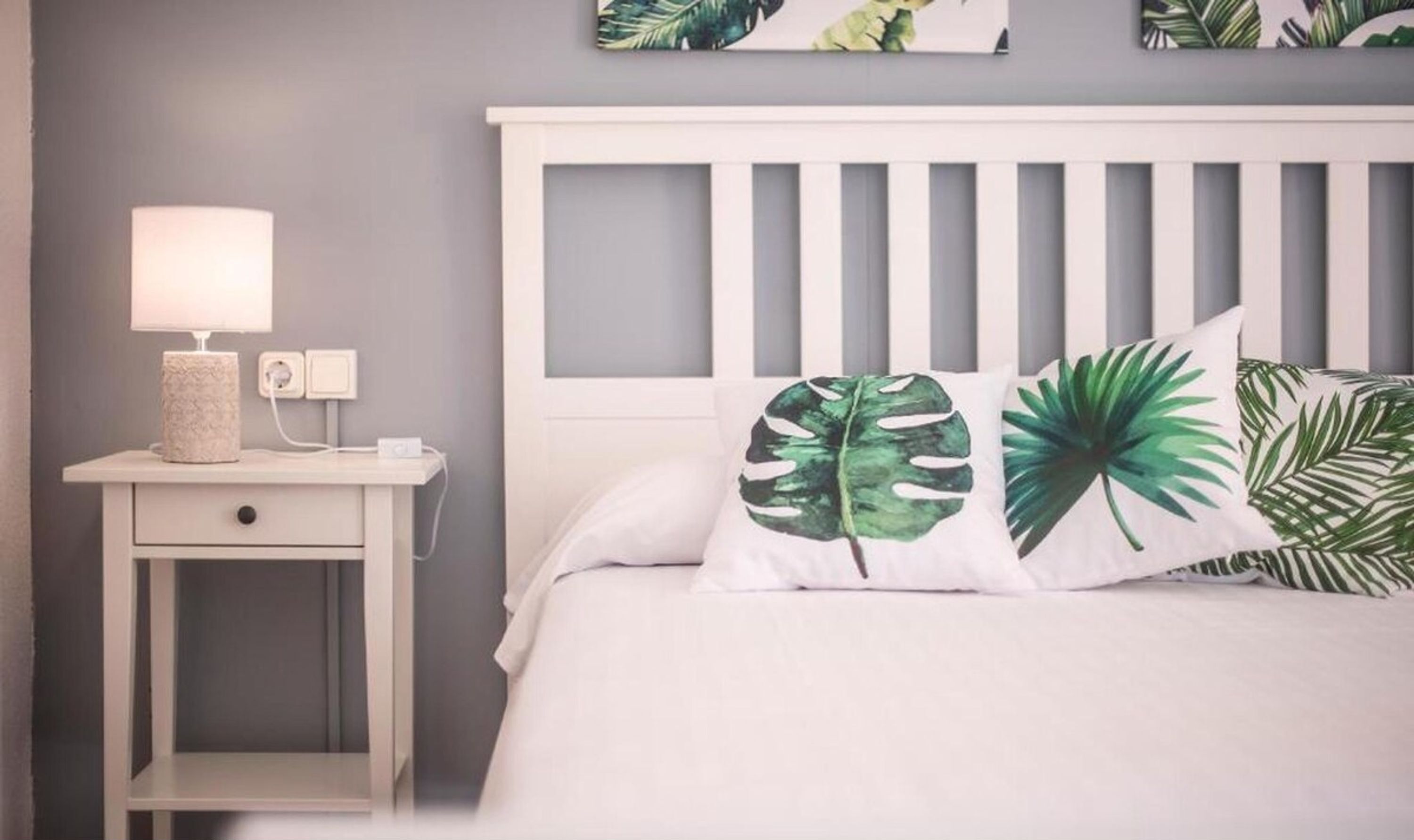 A bedroom with a white bed, two pillows with tropical leaf patterns, a bedside table with a lamp, and a wall-mounted power outlet.