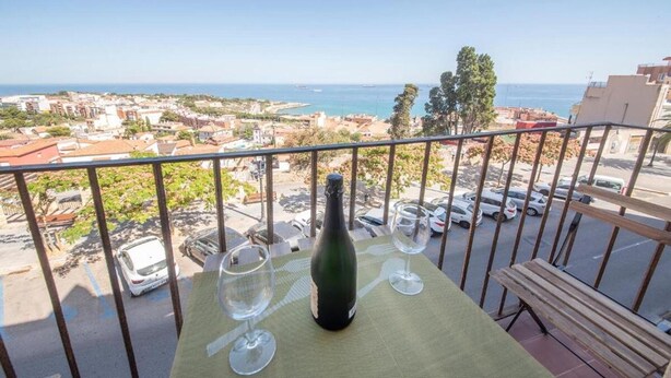 A balcony with a bottle of wine and two glasses on a table, overlooking a coastal cityscape with parked cars and buildings. - Unik Vacation Rovellat (Tarragona)