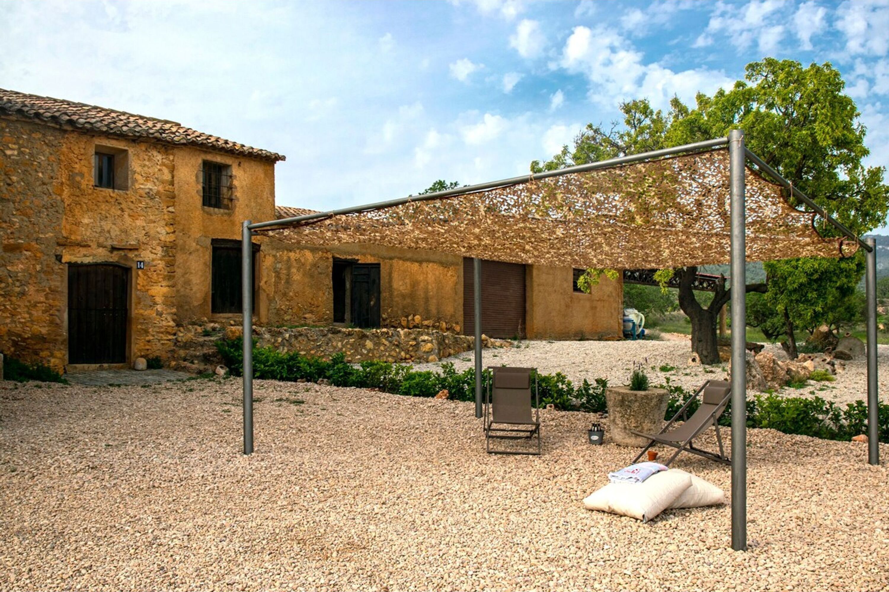 A courtyard with a pergola, two lounge chairs, and a stone building with a tiled roof.