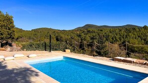 Ein rechteckiger Swimmingpool mit klarem, blauem Wasser, umrahmt von Steinfliesen. Ein Seilzaun ist angebracht und im Hintergrund ist eine bewaldete Bergkette zu sehen.