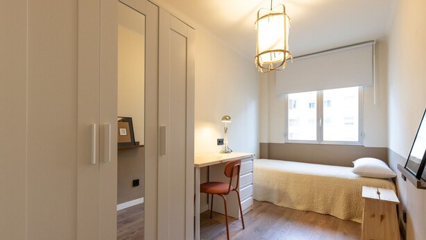 A small bedroom with a bed, a desk, and a chair. There is a window with a view of buildings outside. - Unik Vacation Train Station (Tarragona)