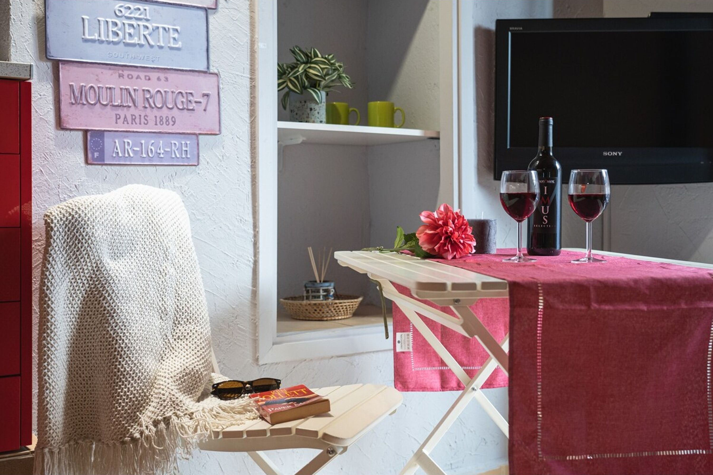 Een gezellige kamer met een witte muur, een rode stoel, een klein tafeltje met een roze tafelkleed, een boekenkast en een televisie.