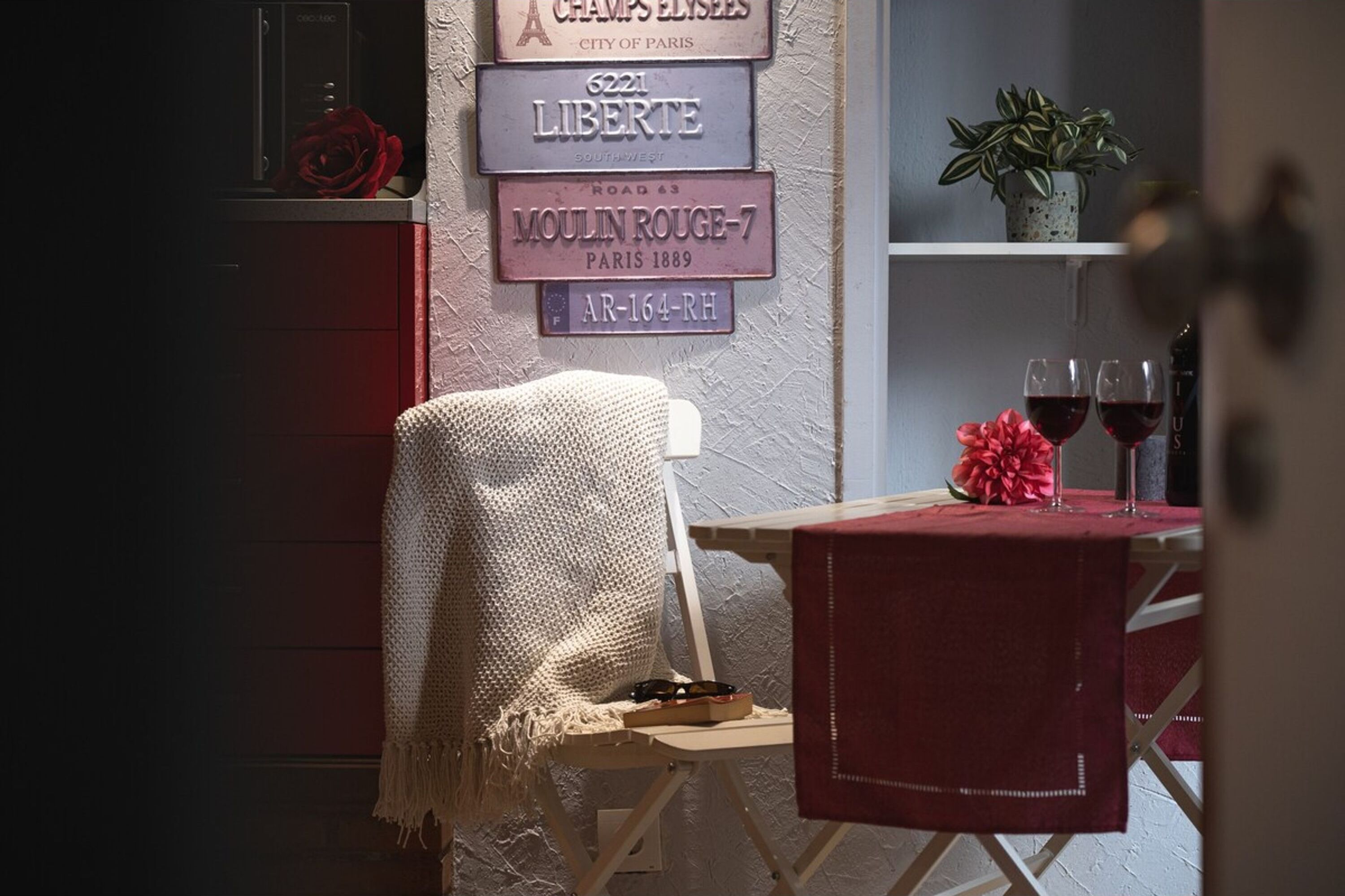 Una habitación acogedora con un letrero antiguo, una pequeña mesa con copas de vino tinto y un sillón con una manta tejida.