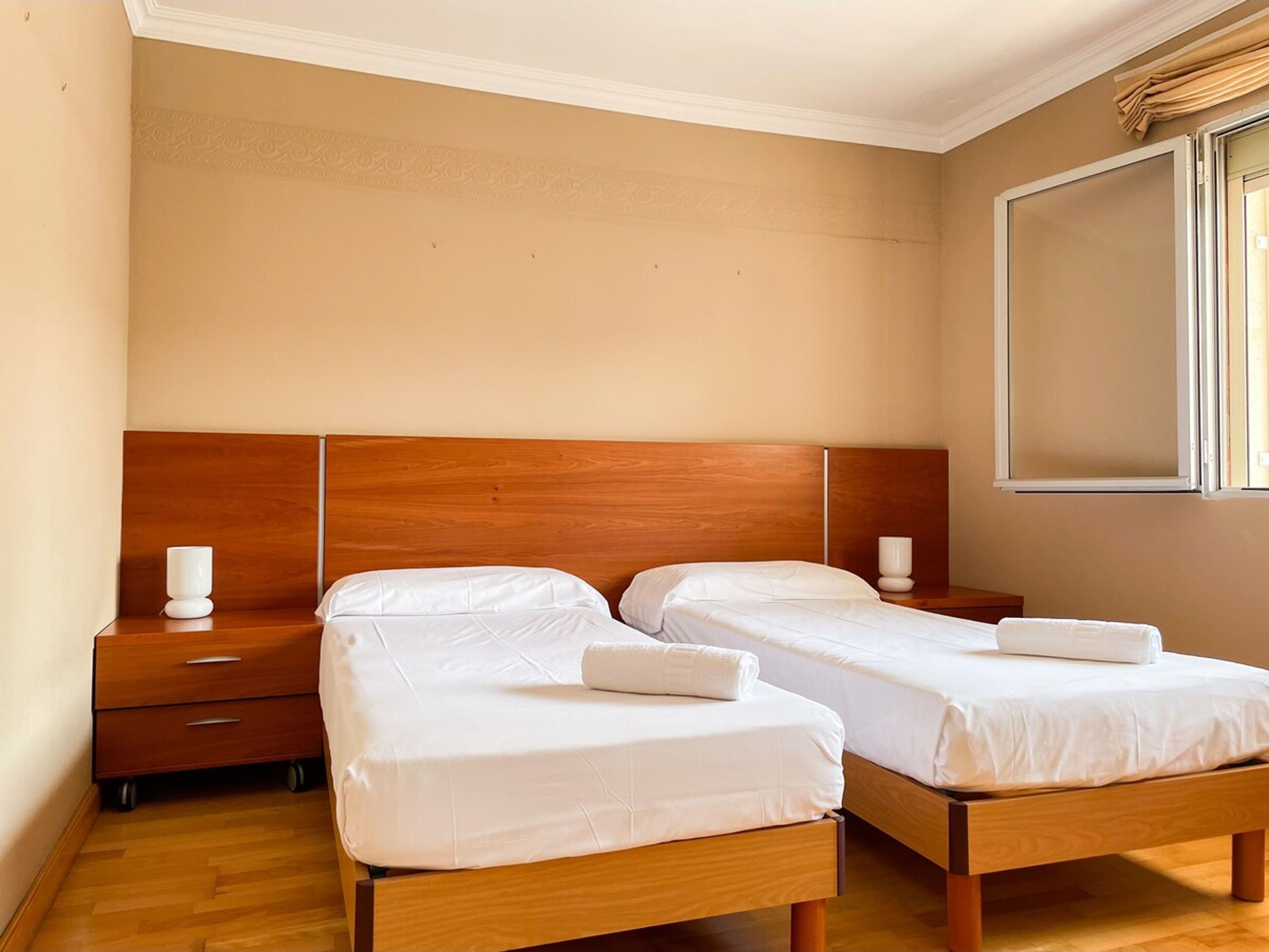 A hotel room with two single beds, wooden headboards, white bedding, a nightstand with a lamp, and a window with curtains.