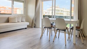 A modern living room with a white sofa, two chairs, a dining table, large windows, and a view of buildings. - Unik Vacation Palm Beach (Vila-Seca)