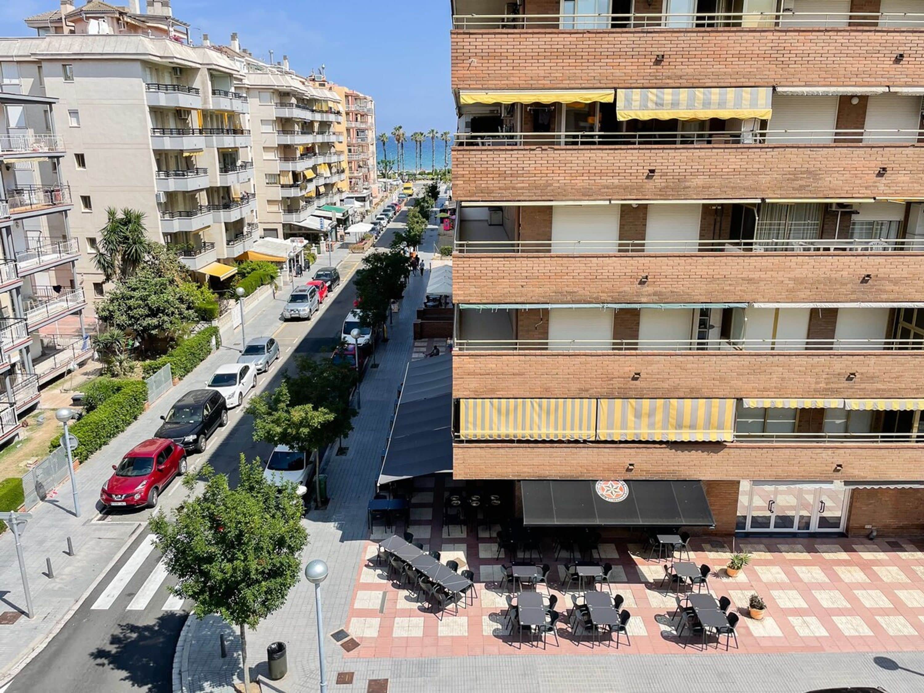 A multi-story building with balconies, a street with parked cars, and outdoor seating.