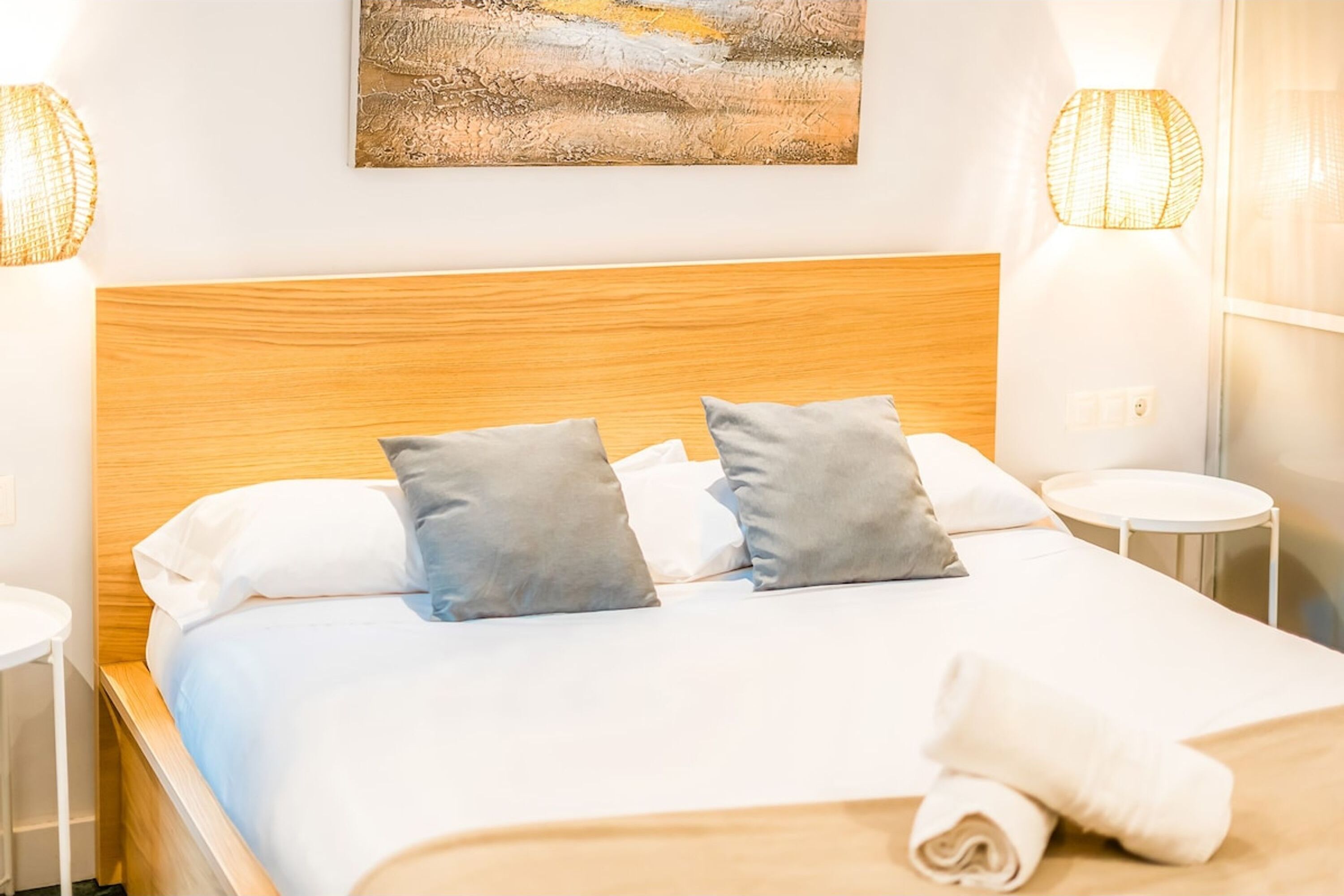 A neatly made bed with a wooden headboard, two gray pillows, and a white blanket. There are two bedside tables with lamps, and a painting is visible on the wall above the bed.