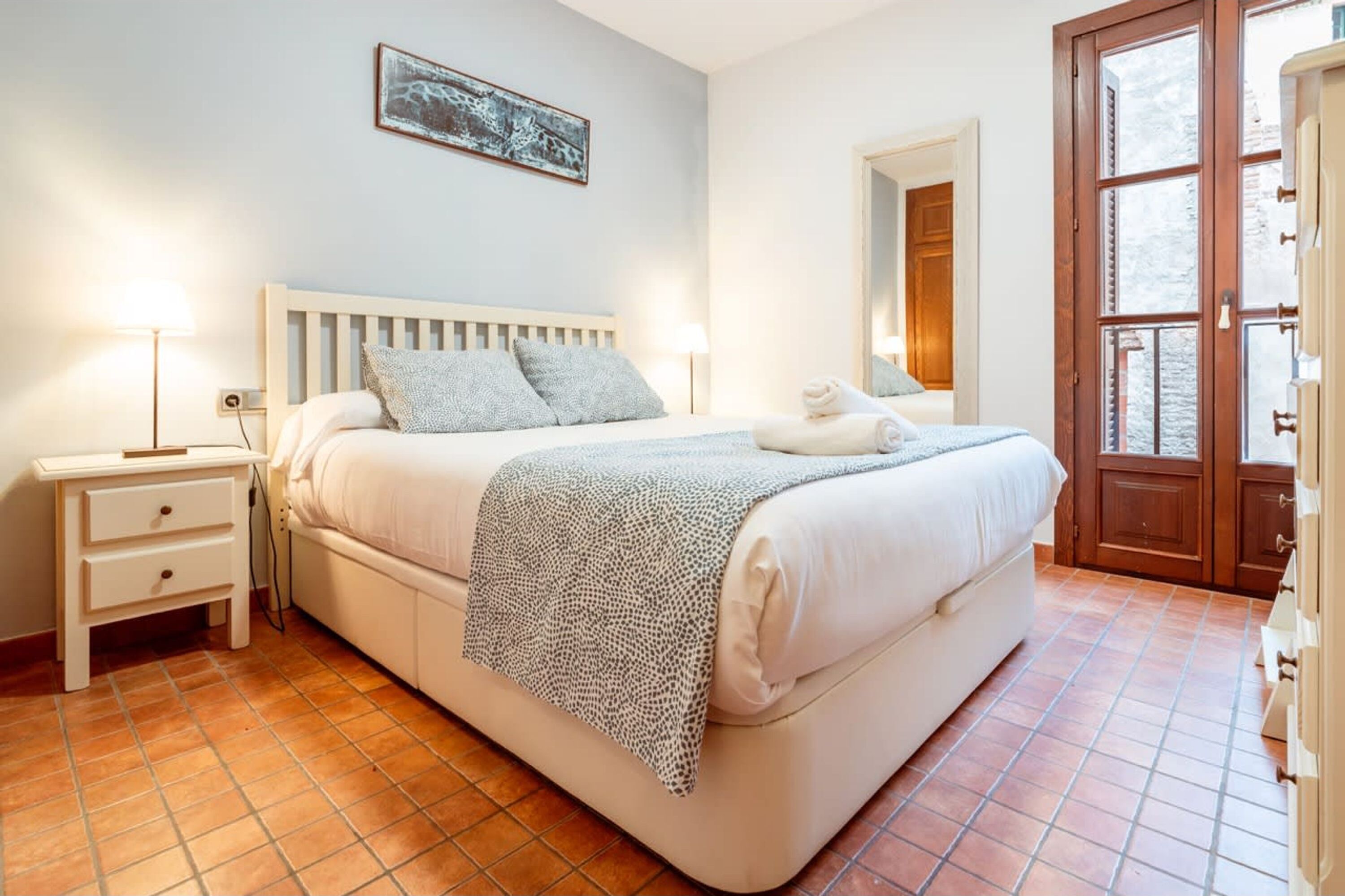 Ein Schlafzimmer mit Bett, Nachttischlampen, einem Nachttisch, einem Wandbild und einer Tür, die auf einen Balkon führt.