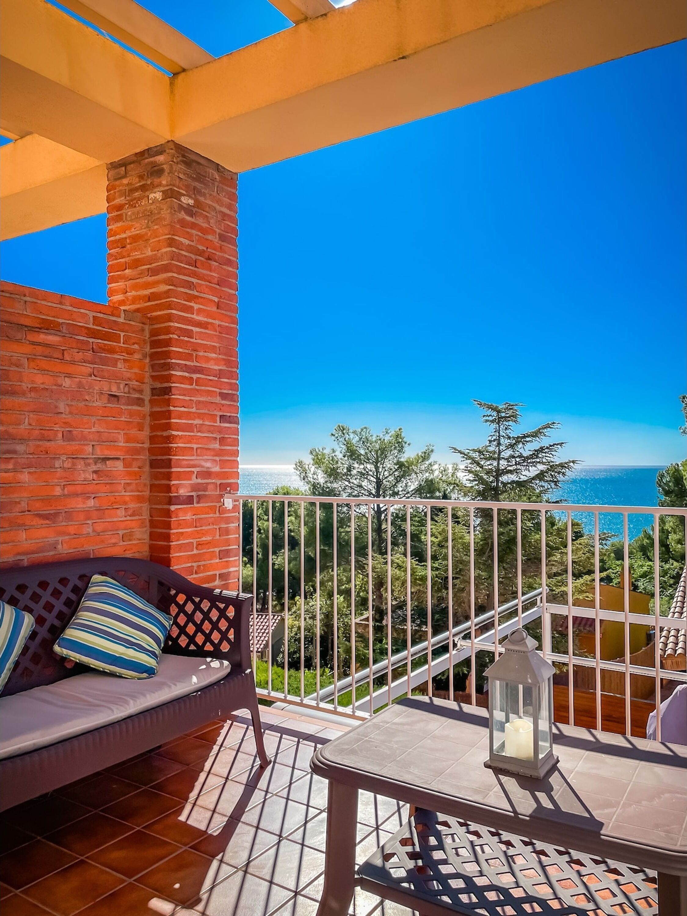 A balcony with a brick wall, a sofa with cushions, a small table with a lantern, and a view of the sea and trees.