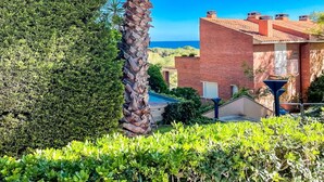 A well-maintained garden with a palm tree, neatly trimmed bushes, and a brick building with a visible garage and outdoor space. - Unik Vacation Tamarit (Tarragona)