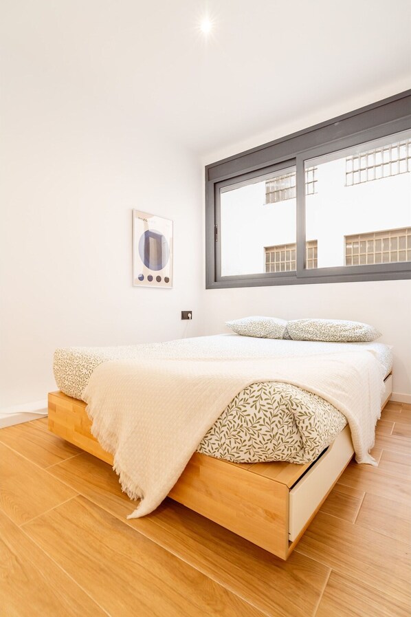 A bedroom with a wooden bed, white bedding, a window with blinds, and a wall-mounted light switch. - Unik Vacation Penthouse (Tarragona)