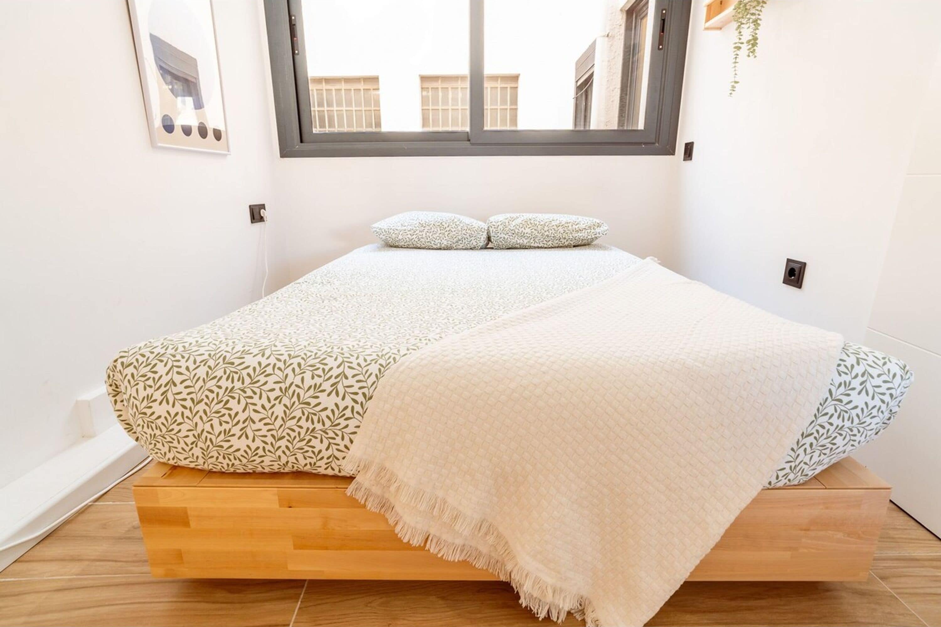 A bed with a patterned pillow and a quilt, a window above, and a plant hanging on the wall.