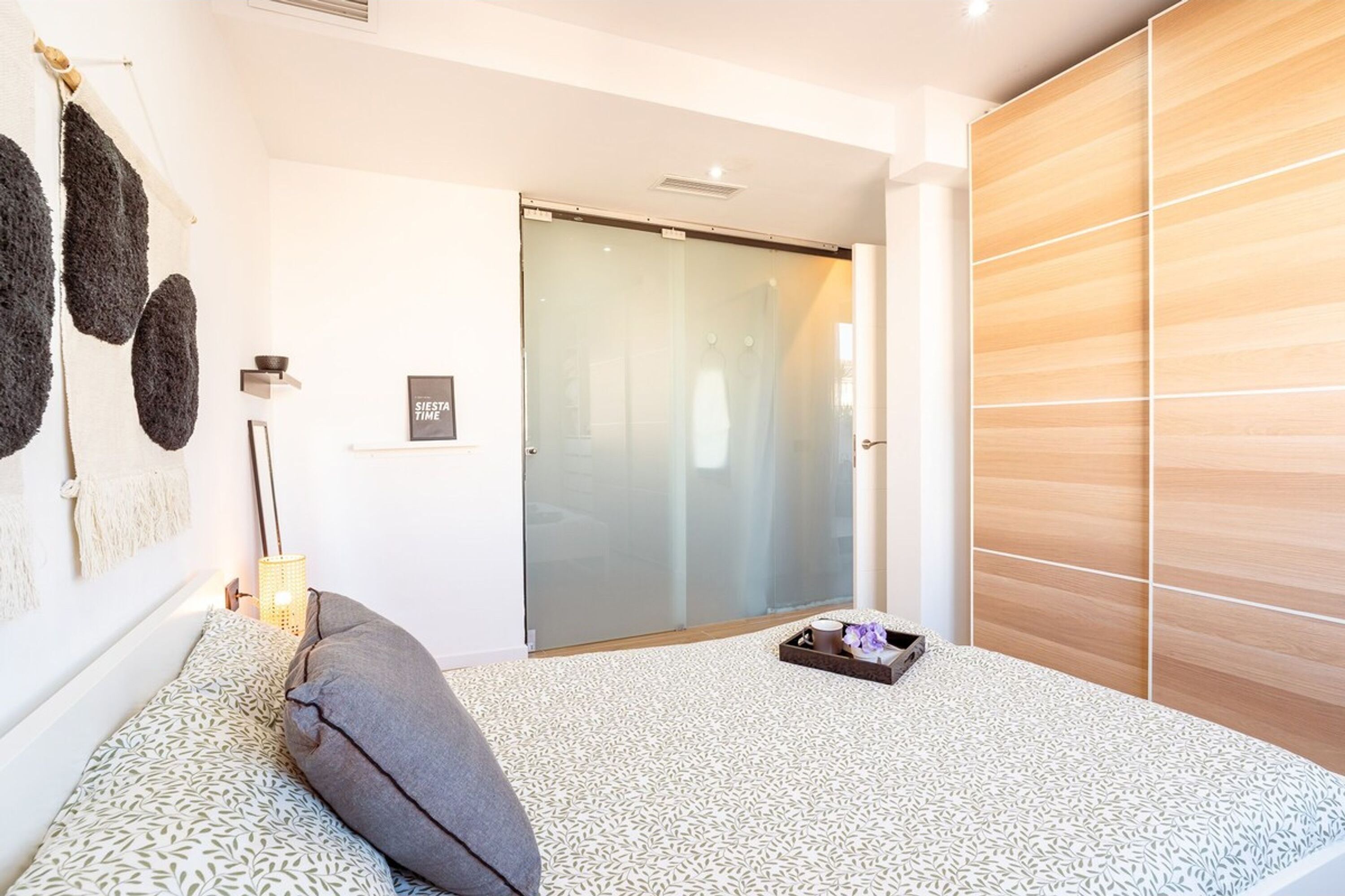 A modern bedroom with a patterned carpet, a bed with a gray pillow, a bedside table with a lamp, a sliding glass door, and a wooden wardrobe.