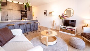 A modern living room with a sofa, coffee table, and bar stools. A TV and lamp are on a stand, and a kitchen area is visible in the background. - Unik Vacation Penthouse (Tarragona)