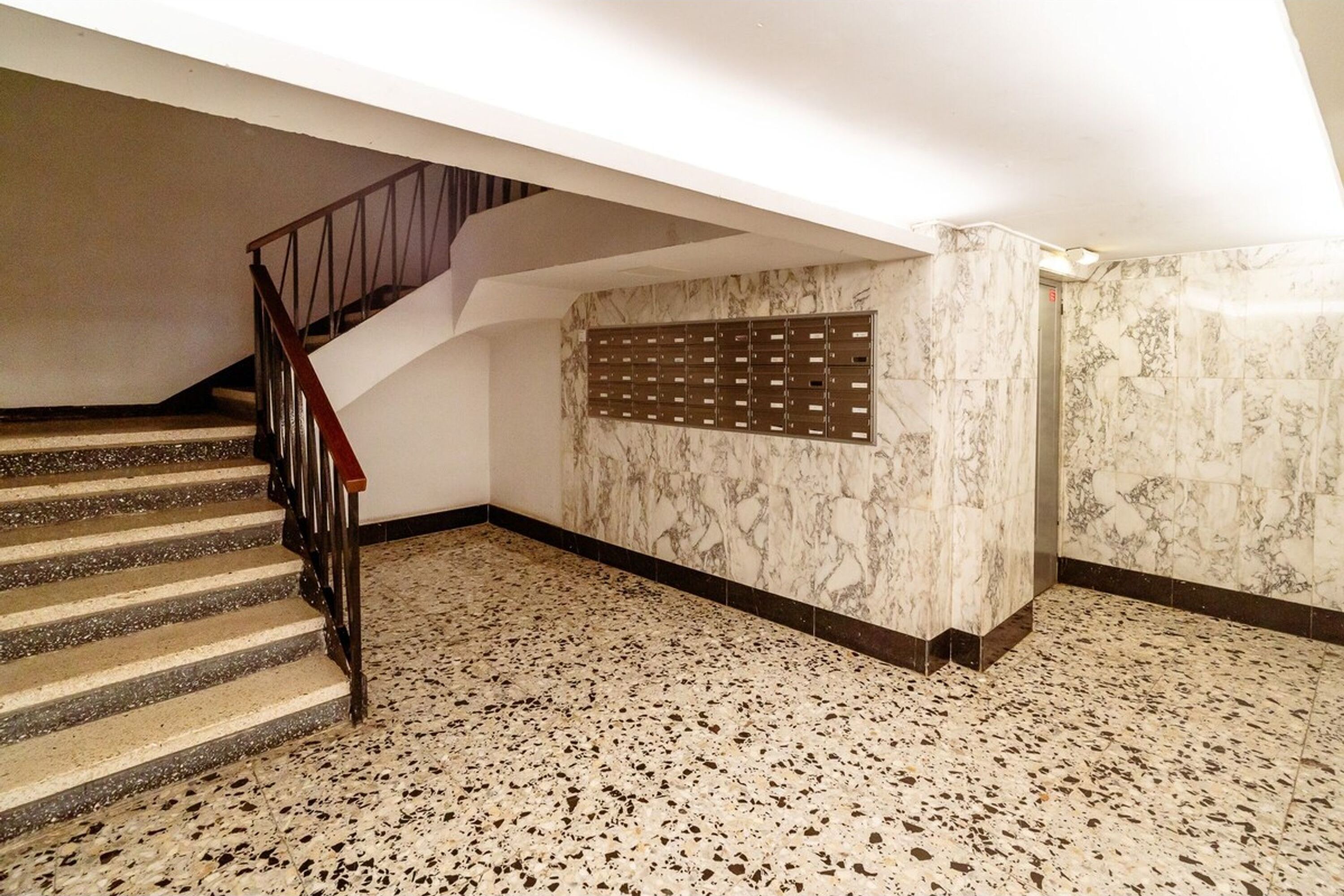 Una escalera con barandilla de madera que conduce a una plataforma. La plataforma tiene una pared de mármol con un panel decorativo. El suelo es moteado.