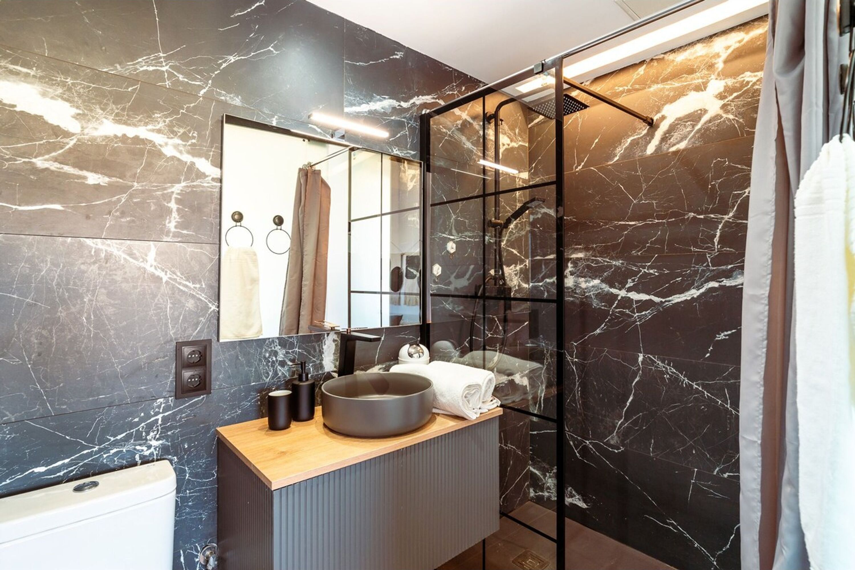 A modern bathroom with a marble backsplash, a wooden vanity, a large mirror, and a glass shower enclosure.