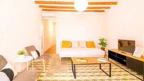 A cozy living room with a white sofa, a coffee table, and a TV. There is a small side table with a plant and a chair. The room has wooden beams on the ceiling and a patterned rug on the floor. - Unik Vacation Congress (Tarragona)