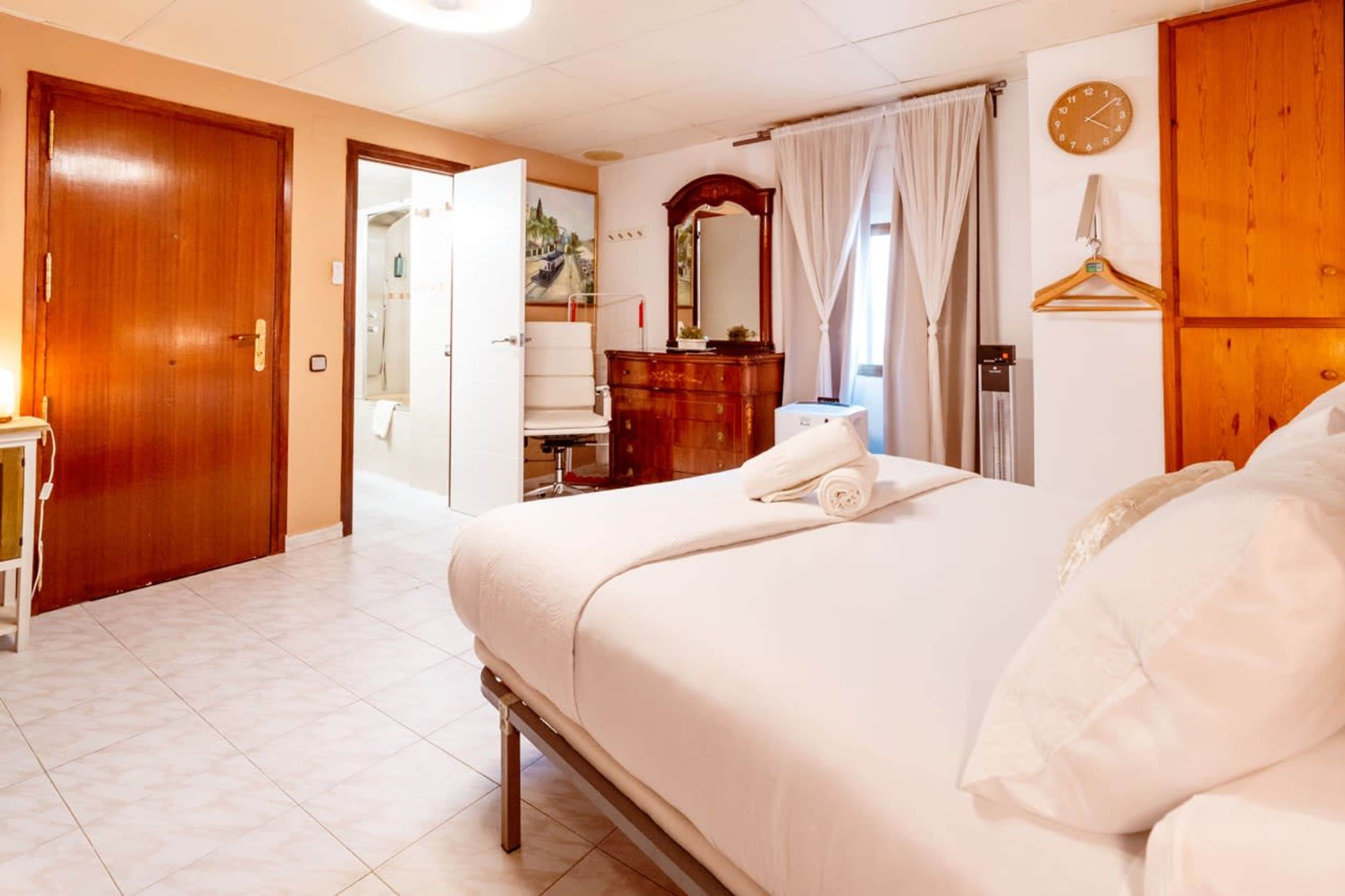 Habitación de hotel con una cama, un tocador de madera, un espejo, un reloj y una puerta que da a un baño.