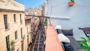 En smal gade med bygninger langs begge sider, en væg med røde teglsten, samt en balkon med en fletsofa og hynder.