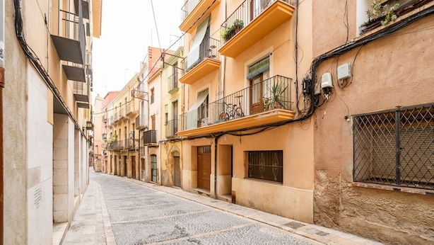 Uma rua estreita ladeada por edifícios altos, com varandas decoradas com plantas em vasos e bicicletas.