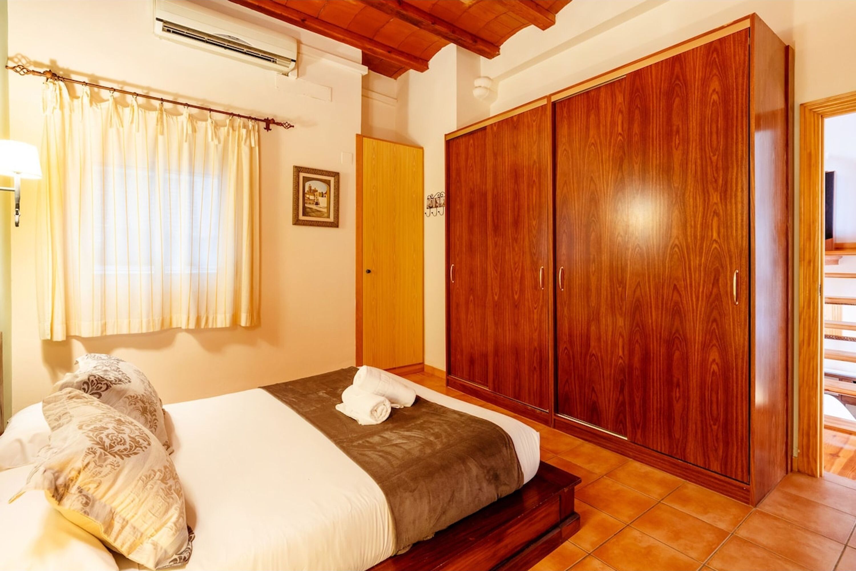 A bedroom with a bed, a wooden wardrobe, a window with curtains, and a picture on the wall.