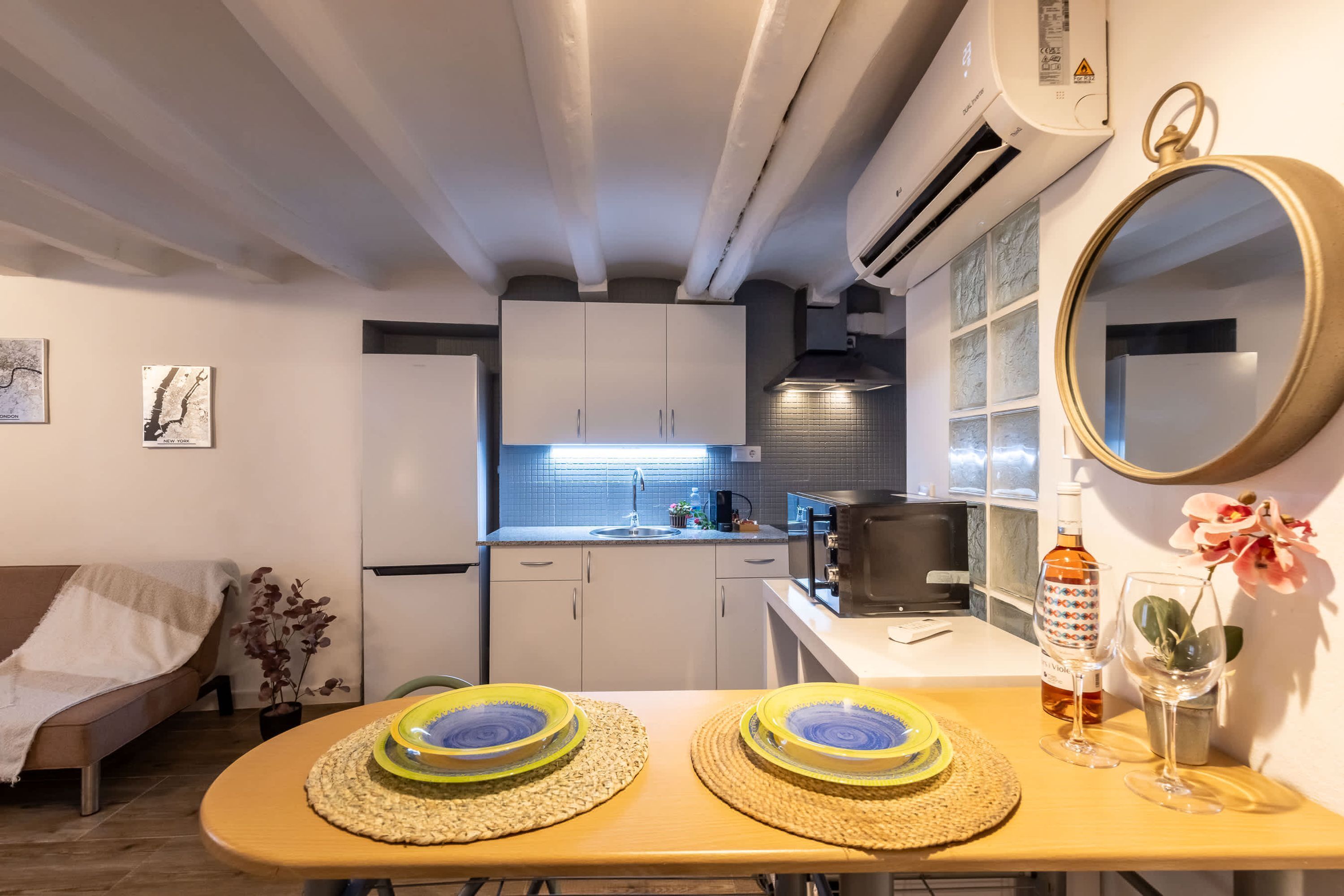 Un espacio habitable compacto con kitchenette, un área de comedor con dos platos, un espejo y una pequeña planta en maceta.