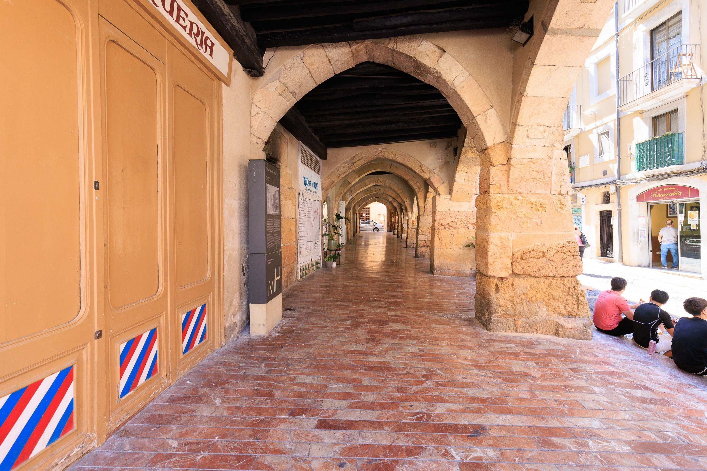 Un couloir bordé d’arches en pierre, doté d’un sol carrelé et d’une rangée de portes sur lesquelles figurent des enseignes de salons de coiffure.