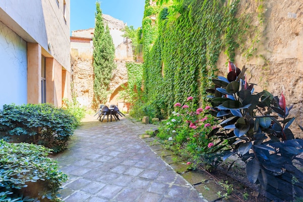 Un étroit sentier de pierre mène à un espace aménagé avec une table et des chaises, entouré d’une végétation luxuriante et d’un mur de pierre.