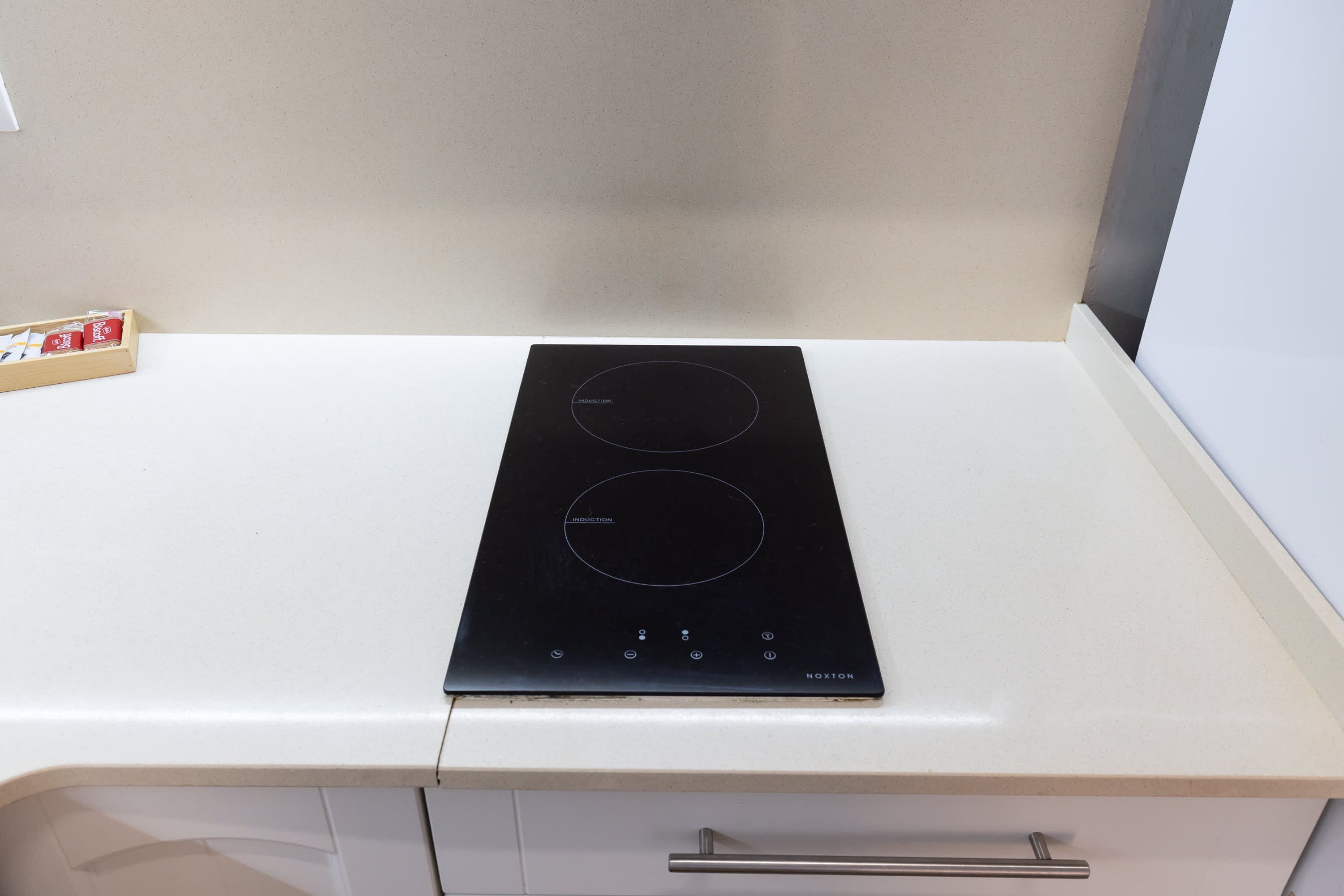 Una cocina de inducción negra sobre una encimera blanca, con una caja de té de madera y una pared blanca al fondo.
