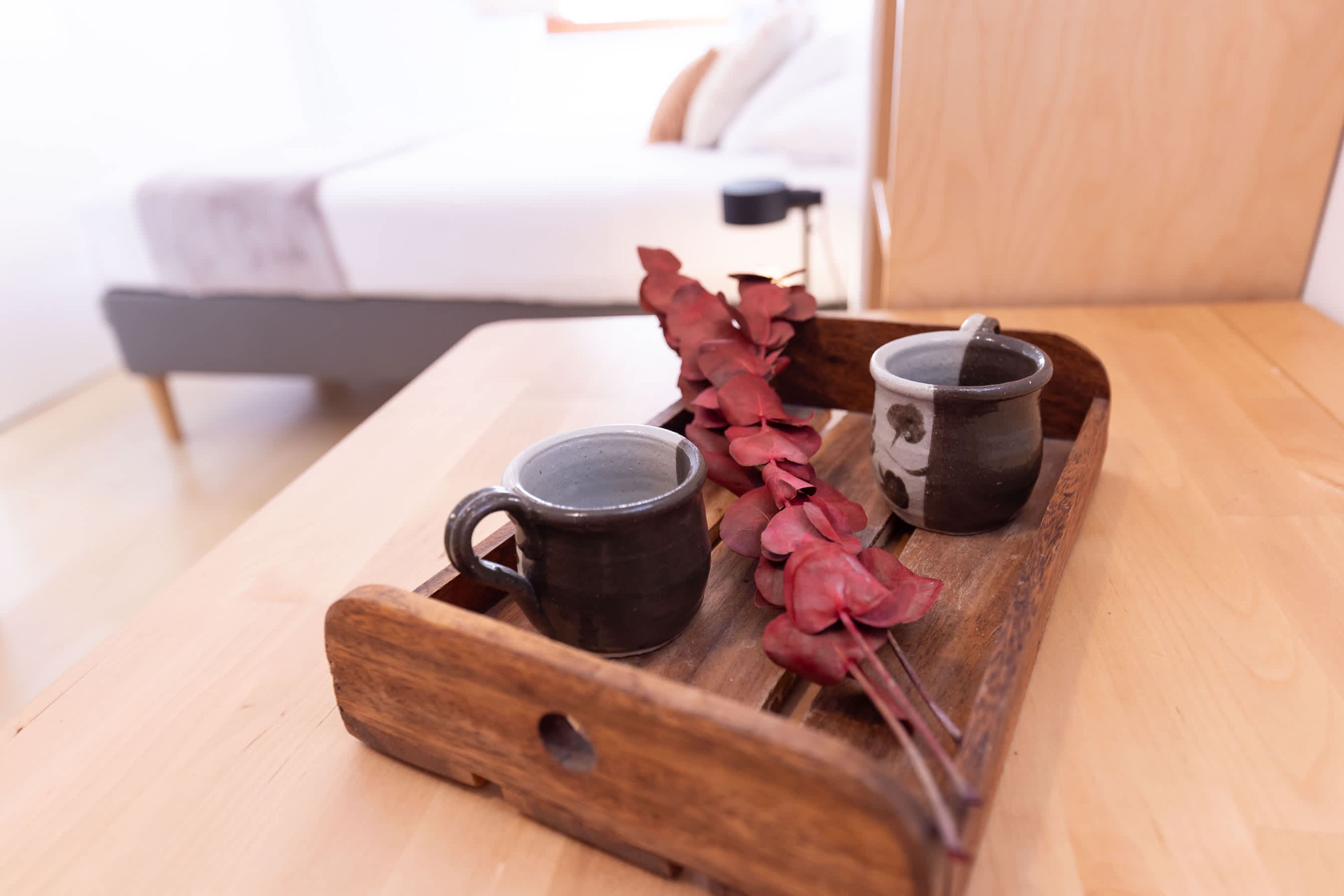 Una bandeja de madera con dos tazas y hojas rojas secas sobre una mesa en una habitación con una cama y una mesita de noche.