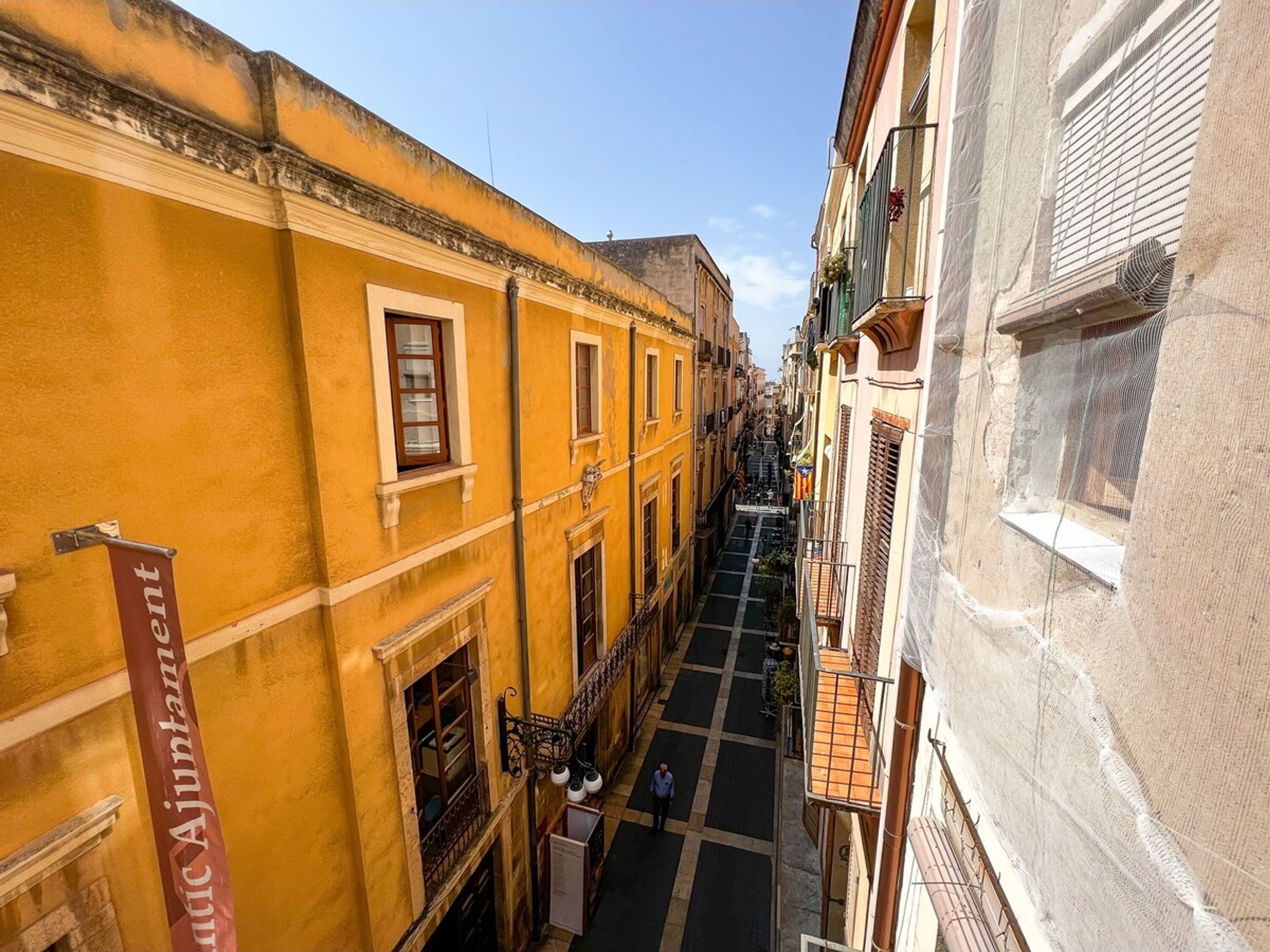 A narrow street with yellow buildings, a sign reading "Hotel Ajuntam", and a person walking.