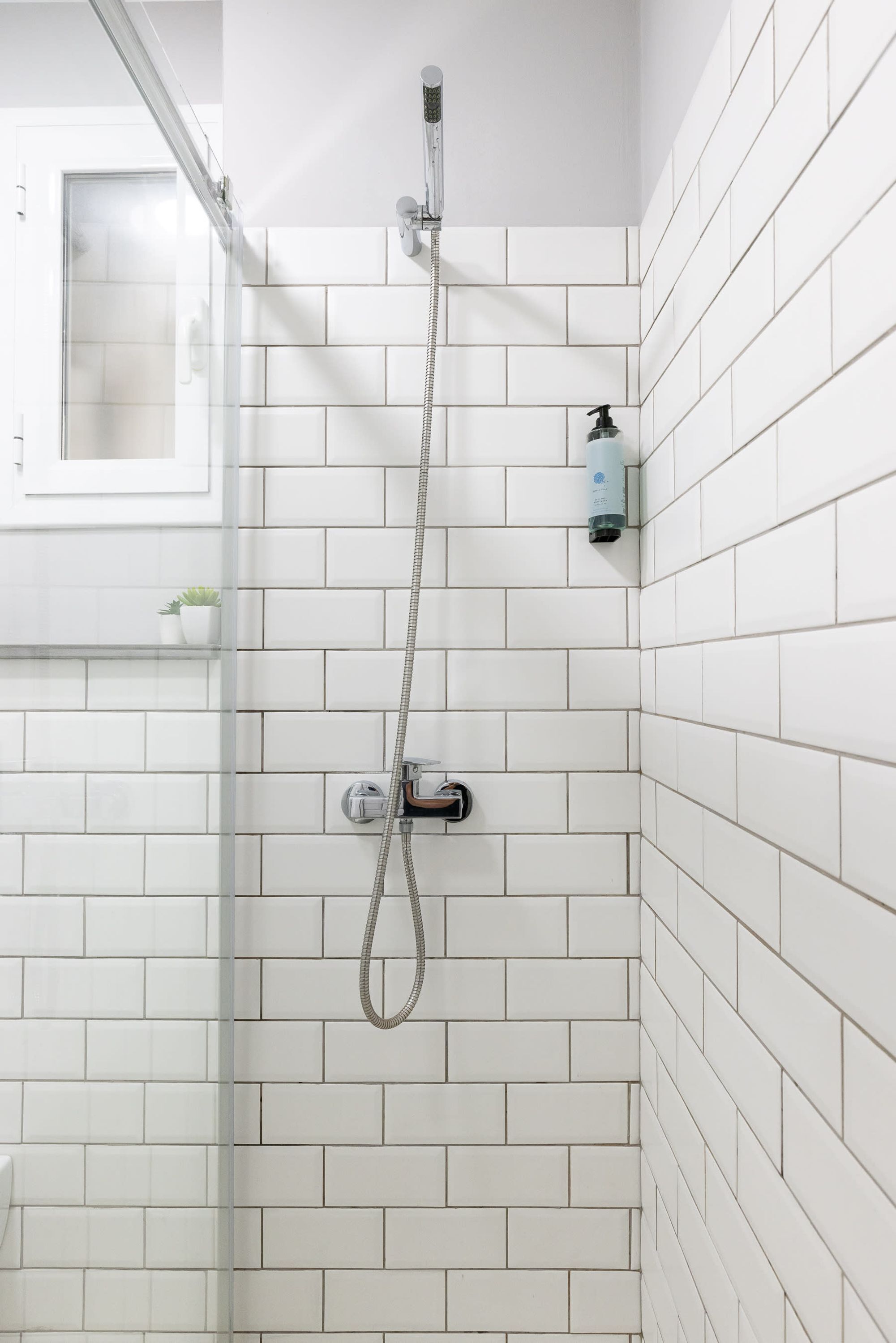 A shower with a glass door, white tiled walls, and a small shelf with a plant.