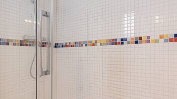 A glass-enclosed shower with a handheld showerhead, a white tiled wall with a colorful mosaic border, and a small white sink with a bottle of hand soap.