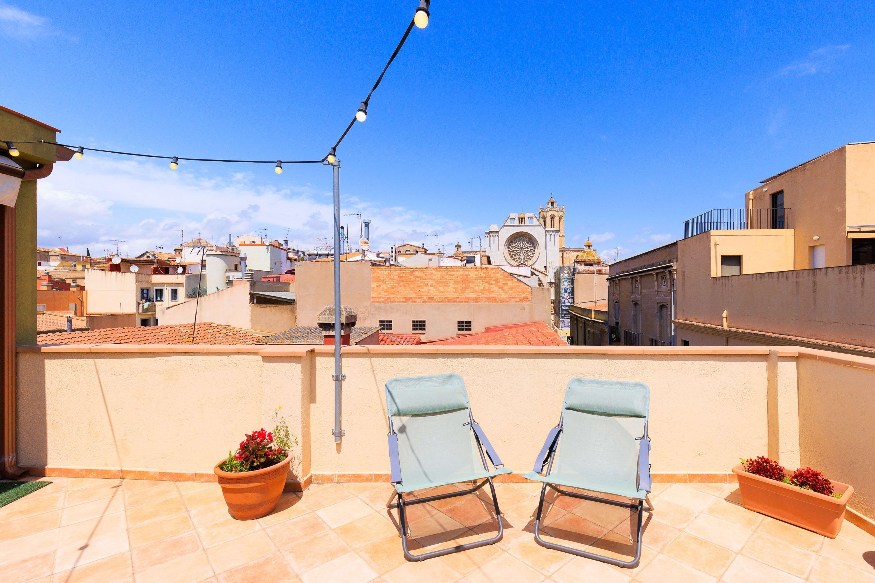 A rooftop terrace with two lounge chairs, potted plants, and a string of lights against a cityscape backdrop.