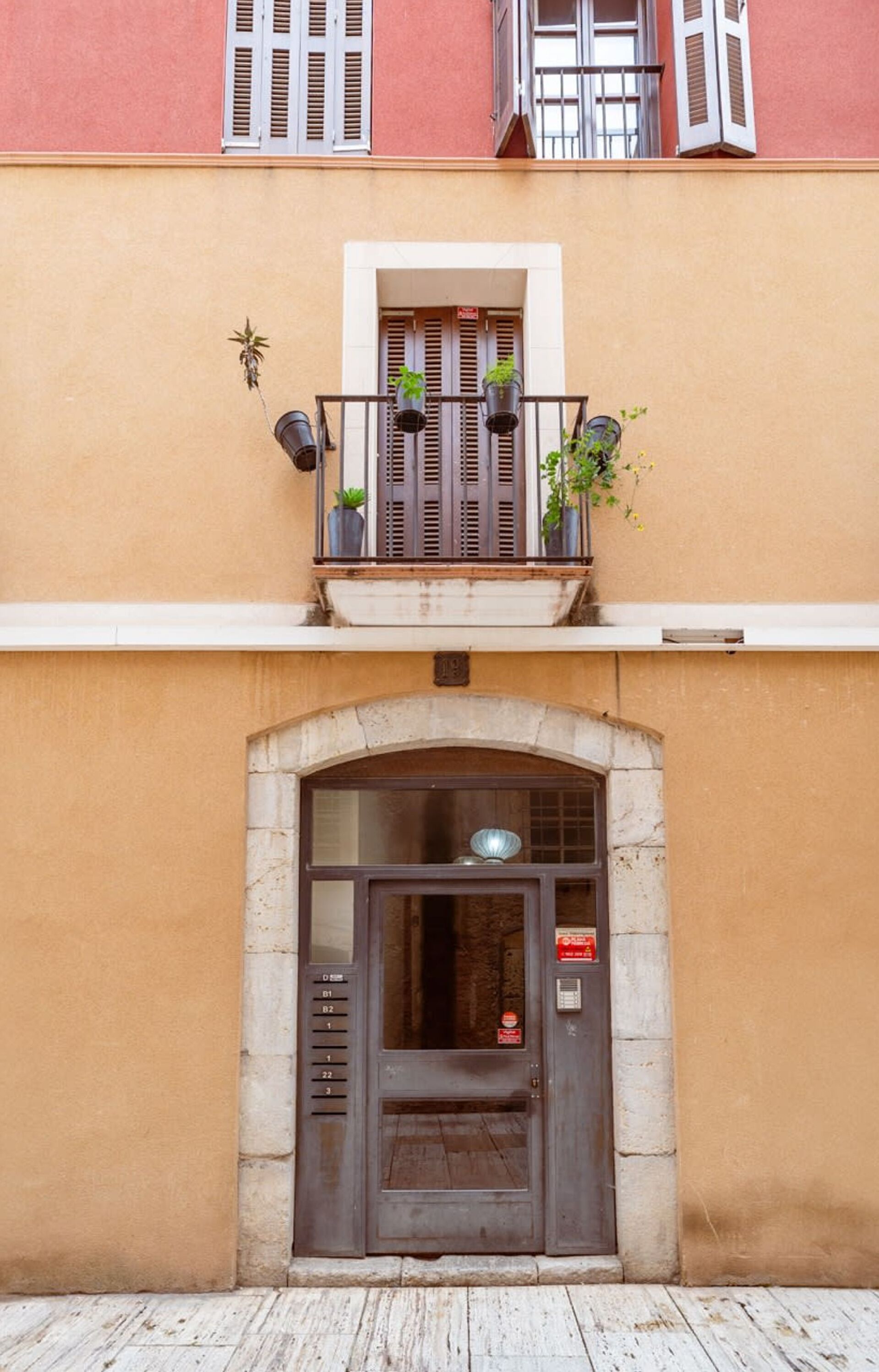 Un edificio con entrada a nivel de calle, un pequeño balcón con plantas en macetas y una ventana con persianas.